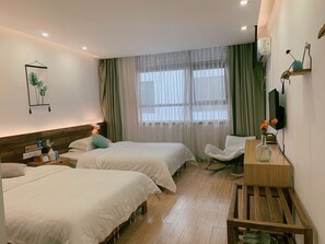 Habitación de hotel con dos camas, cabecera de madera, mesita de noche con lámpara, una silla, escritorio con un florero y ventana con cortinas traslúcidas.