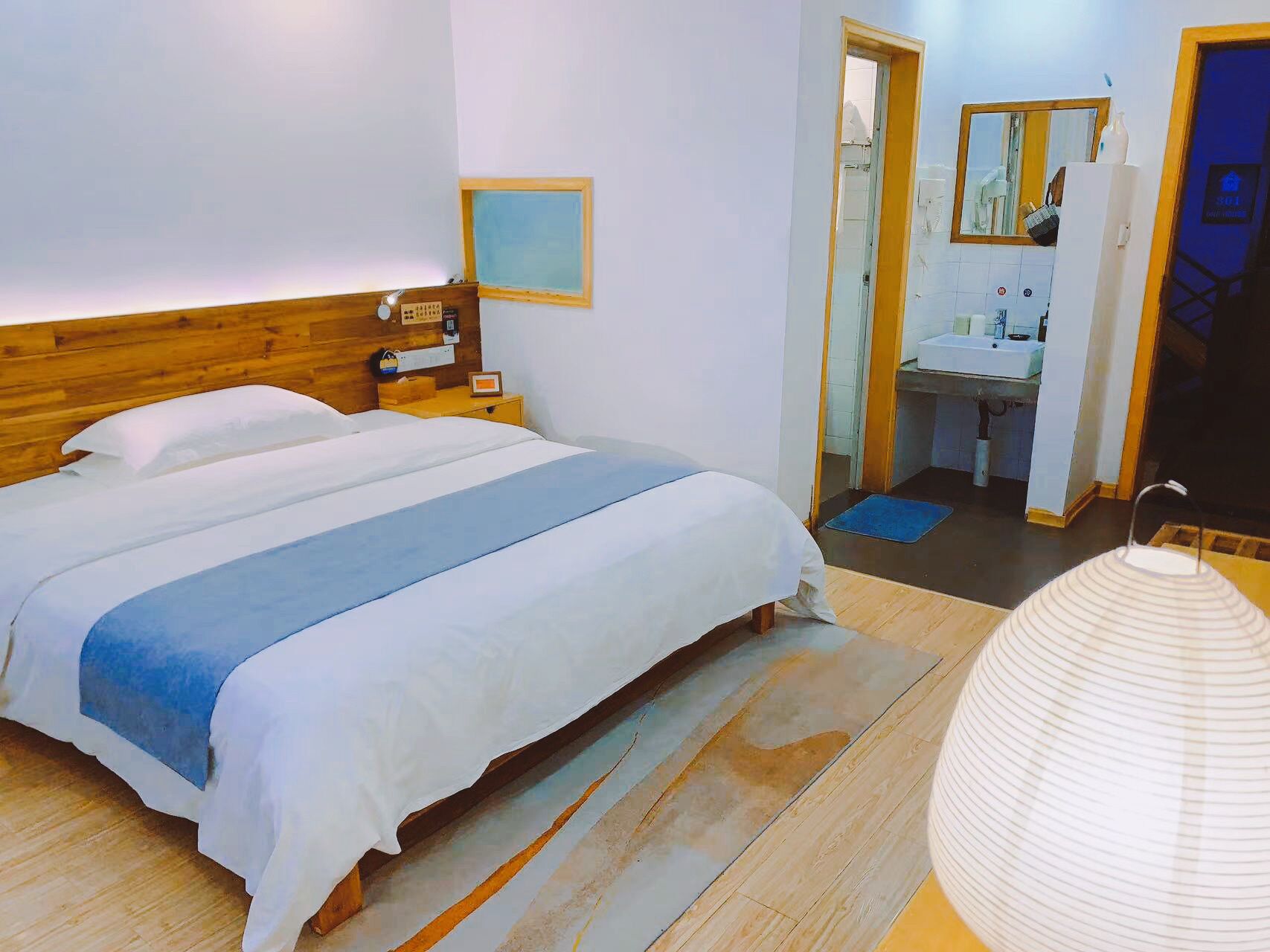 Une chambre à coucher avec une tête de lit en bois, un lit avec une literie blanche et bleue, une table de chevet avec une lampe, un petit tableau encadré et une commode. On aperçoit une salle de bain à travers une porte ouverte, avec un lavabo, un miroir et un porte-serviettes.