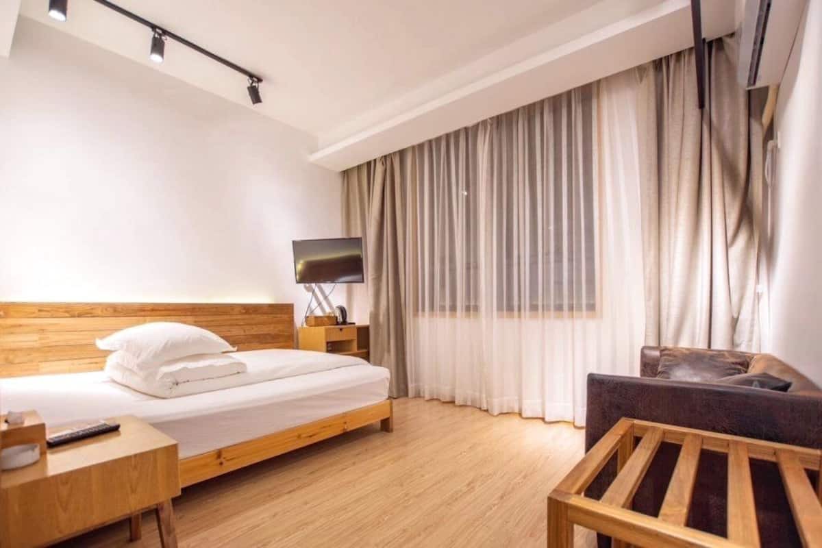 Habitación de hotel con cama de madera, televisor, mesita de noche de madera, silla de madera y una ventana grande con cortinas.