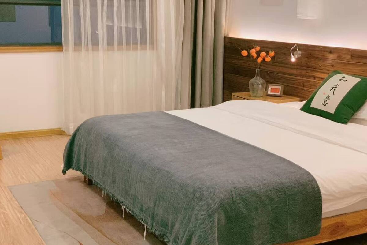 Una cama bien tendida con una manta verde, un cabecero de madera y una mesita de noche con un jarrón y un reloj.