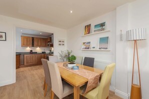 A dining area with a wooden table, chairs, and a potted plant. There is a kitchen in the background with wooden cabinets and a countertop. - Host & Stay | Sunset (Newquay)