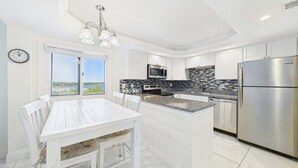 Una cocina moderna con armarios blancos, electrodomésticos de acero inoxidable, encimera de granito y comedor con mesa y sillas blancas.