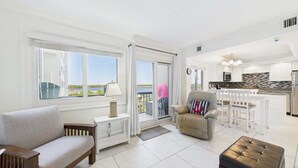 Amplia sala de estar con sofá, sillón y reposapiés, juego de comedor y cocina con armarios blancos y salpicadero negro.