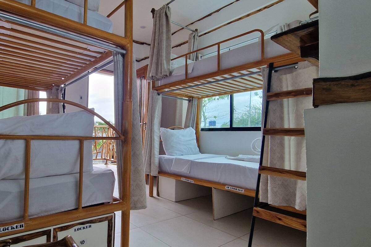 A dormitory room with bunk beds, a window with curtains, and a view of the outdoors.