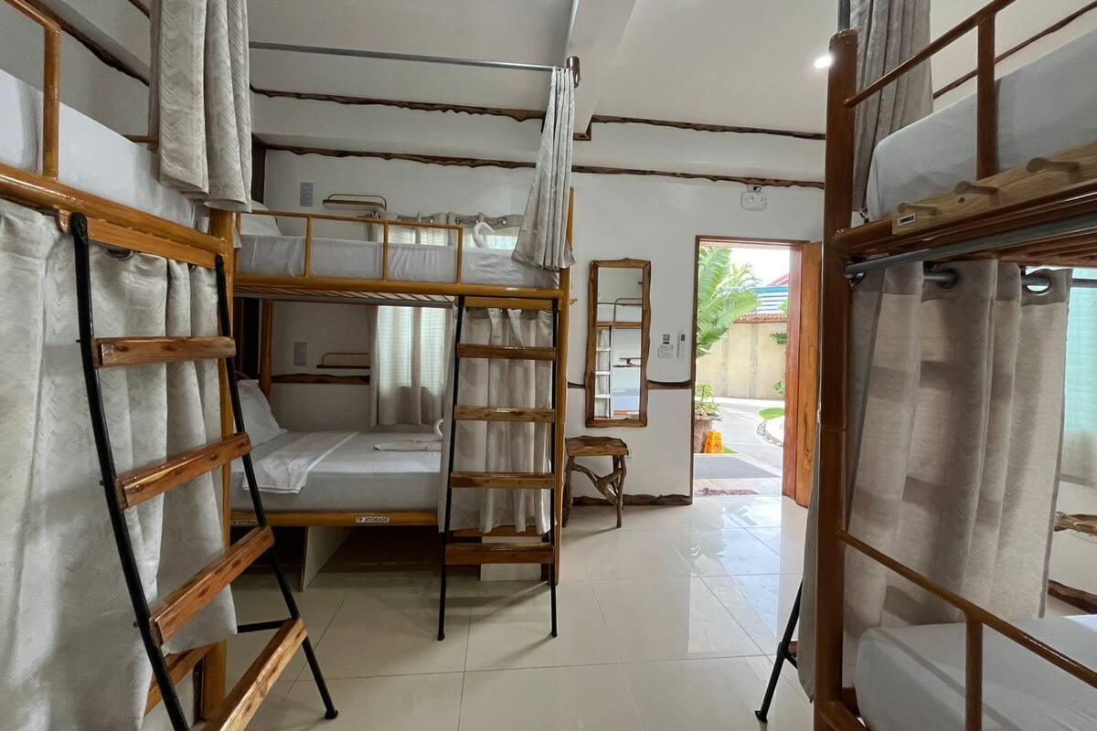 A dormitory room with bunk beds, a small table, a mirror, and a door leading to an outdoor area.