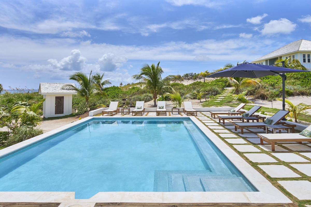 Ein Swimmingpool mit Liegestühlen und Sonnenschirmen, umgeben von viel Grün und einem Haus im Hintergrund.