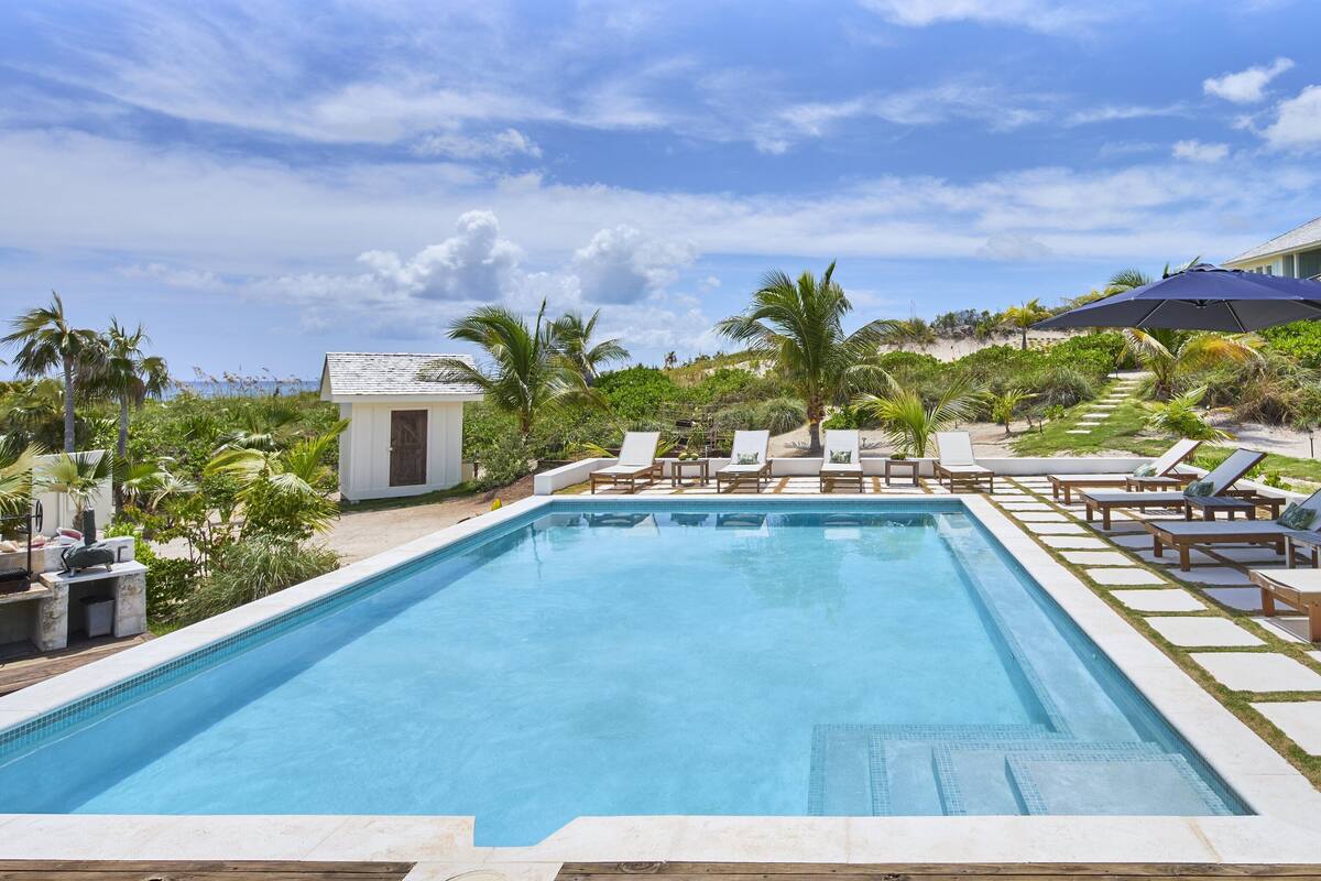 A swimming pool with lounge chairs and umbrellas, surrounded by palm trees and a small building.