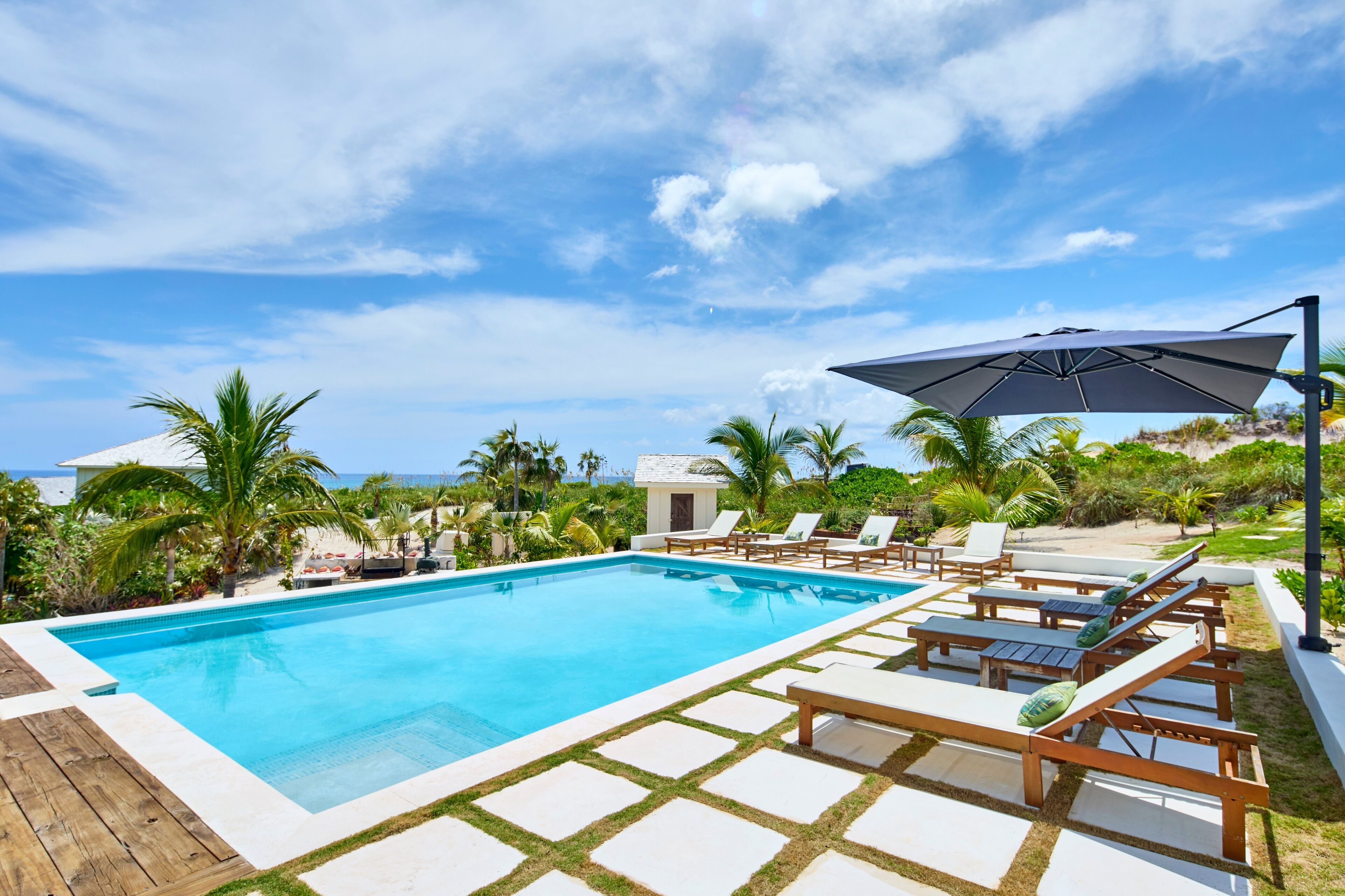 A swimming pool with lounge chairs and an umbrella, surrounded by palm trees and a view of the ocean.
