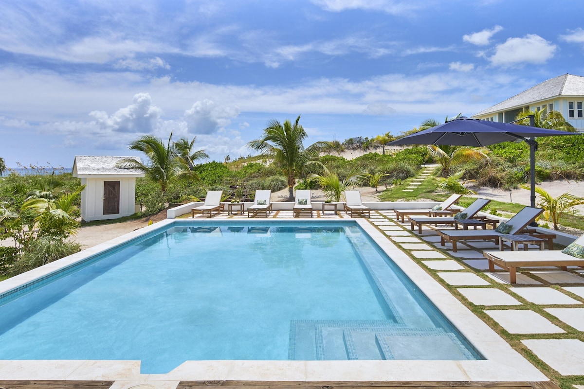 A swimming pool with lounge chairs and umbrellas, surrounded by palm trees and a small building.