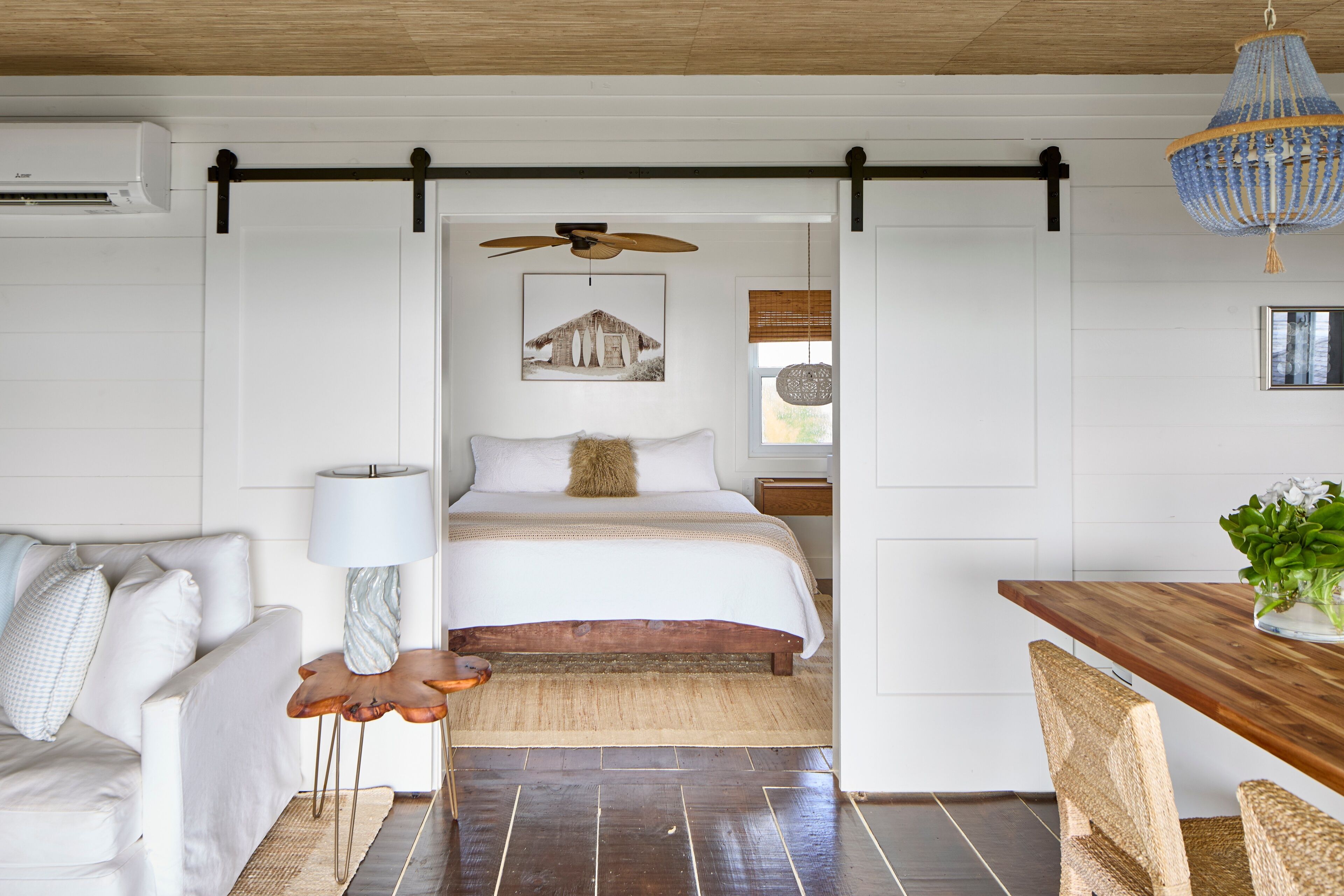 Ein Schlafzimmer mit einem Bett, Holzboden, weißer Wand, Deckenventilator und einem Essbereich mit Tisch und Stühlen.