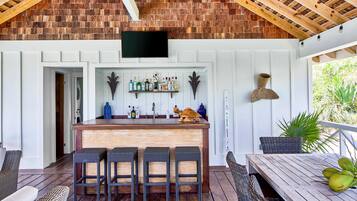 A covered outdoor bar area with a wooden ceiling, a flat-screen TV, a bar counter with stools, and a dining table with a plant.