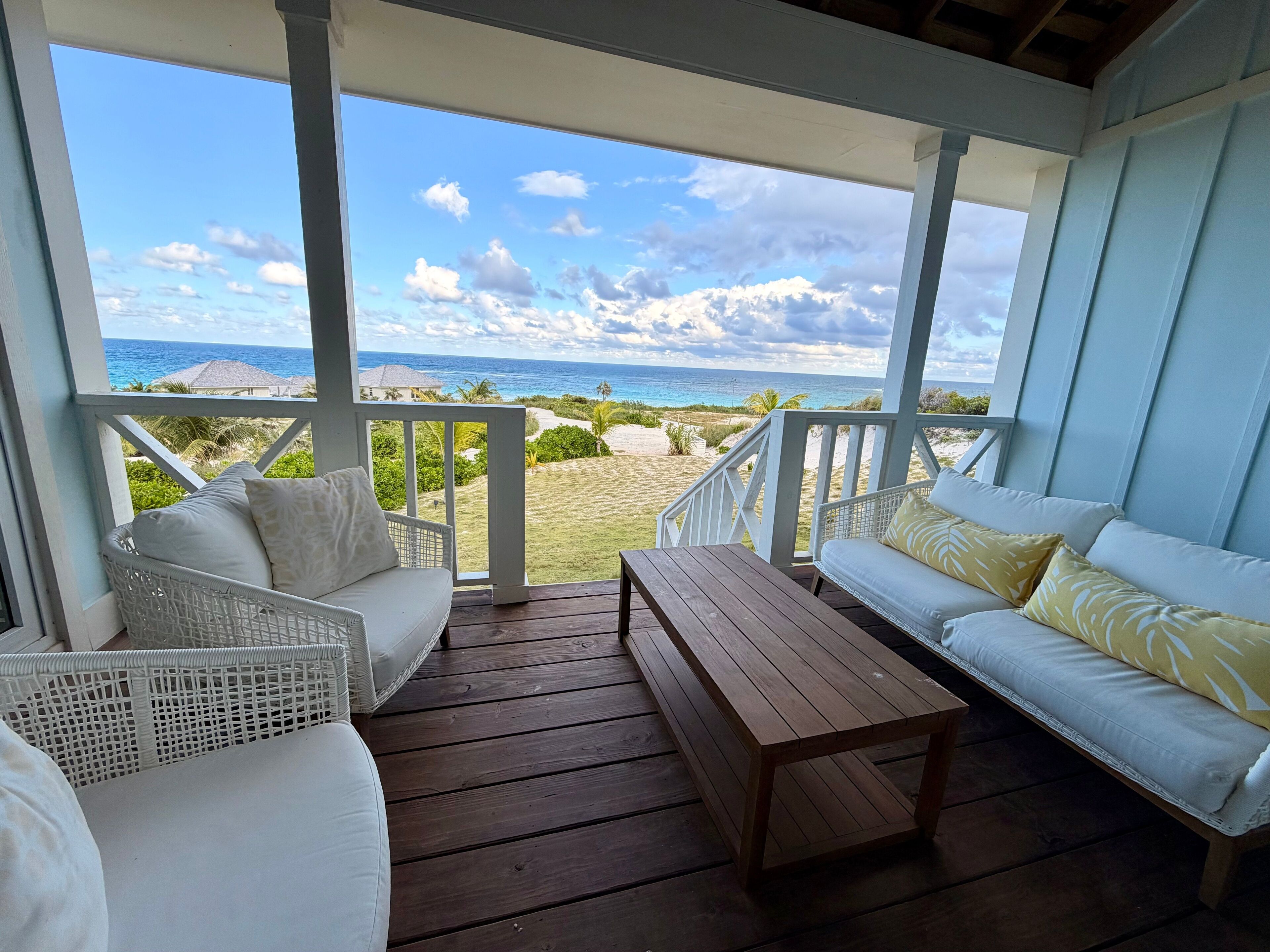 A covered outdoor seating area with white wicker furniture and a wooden table, overlooking a beach with palm trees and the ocean.