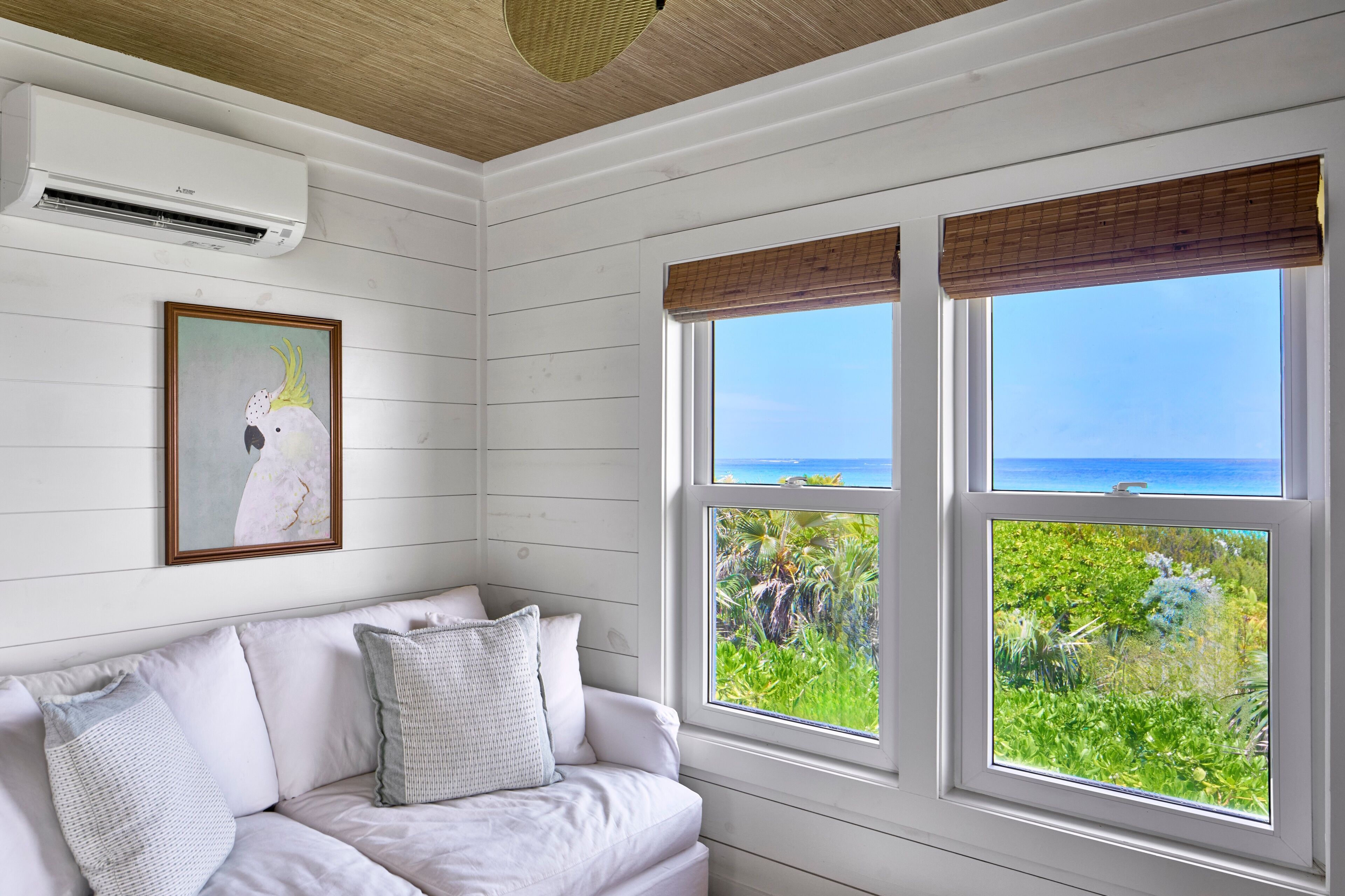 A room with a white couch, a framed artwork, and large windows with a view of greenery and the sea.