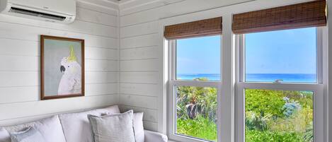 A room with a white couch, a framed artwork, and large windows with a view of greenery and the sea.