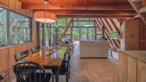 A rustic wooden interior with a dining table, chairs, a sofa, and large windows offering a view of the outdoors.