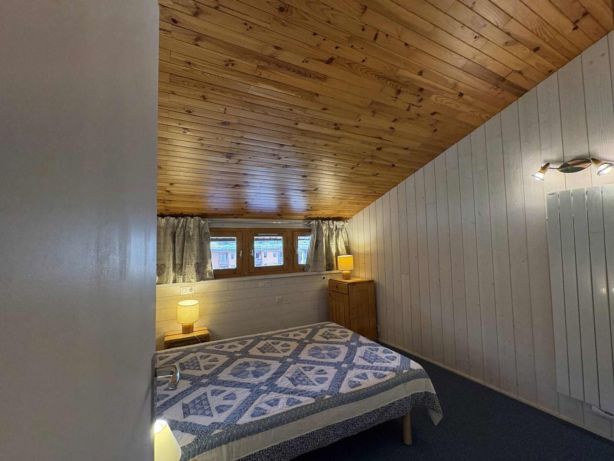 Une chambre à coucher avec un plafond en bois, un lit recouvert d’une couvre-lit à motifs, deux lampes de chevet et une fenêtre avec des rideaux.