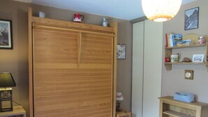 A room with a wooden wardrobe, a small table with a lamp, a shelf with books and decorative items, and a wall clock.