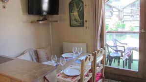 A dining area with a table set for two, a television above the table, and a window with a view of the outside.
