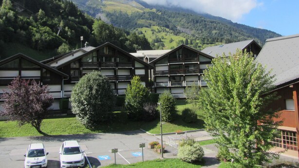 A hotel with traditional wooden architecture, a parking area, and a mountainous backdrop.