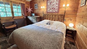 A cozy wooden cabin bedroom with a bed, dresser, vanity, and a window with curtains.