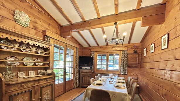 A wooden interior with a dining table set for four, surrounded by chairs. There are shelves with decorative plates and a cabinet with intricate designs.