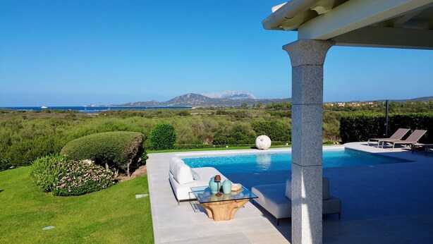 A pool area with a glass table, lounge chairs, and a view of the sea and mountains. - Villa Blue Sphere by Wonderful Italy (Olbia)