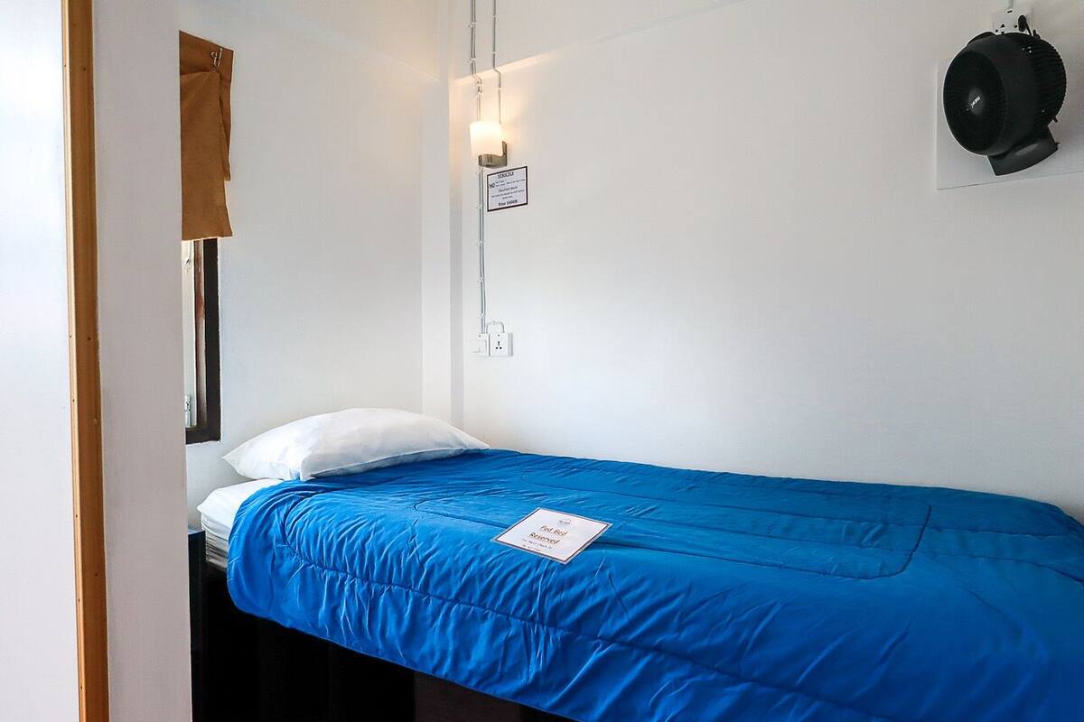 A single bed with a blue mattress and white pillows, a wall-mounted light fixture, and a black speaker on the wall.