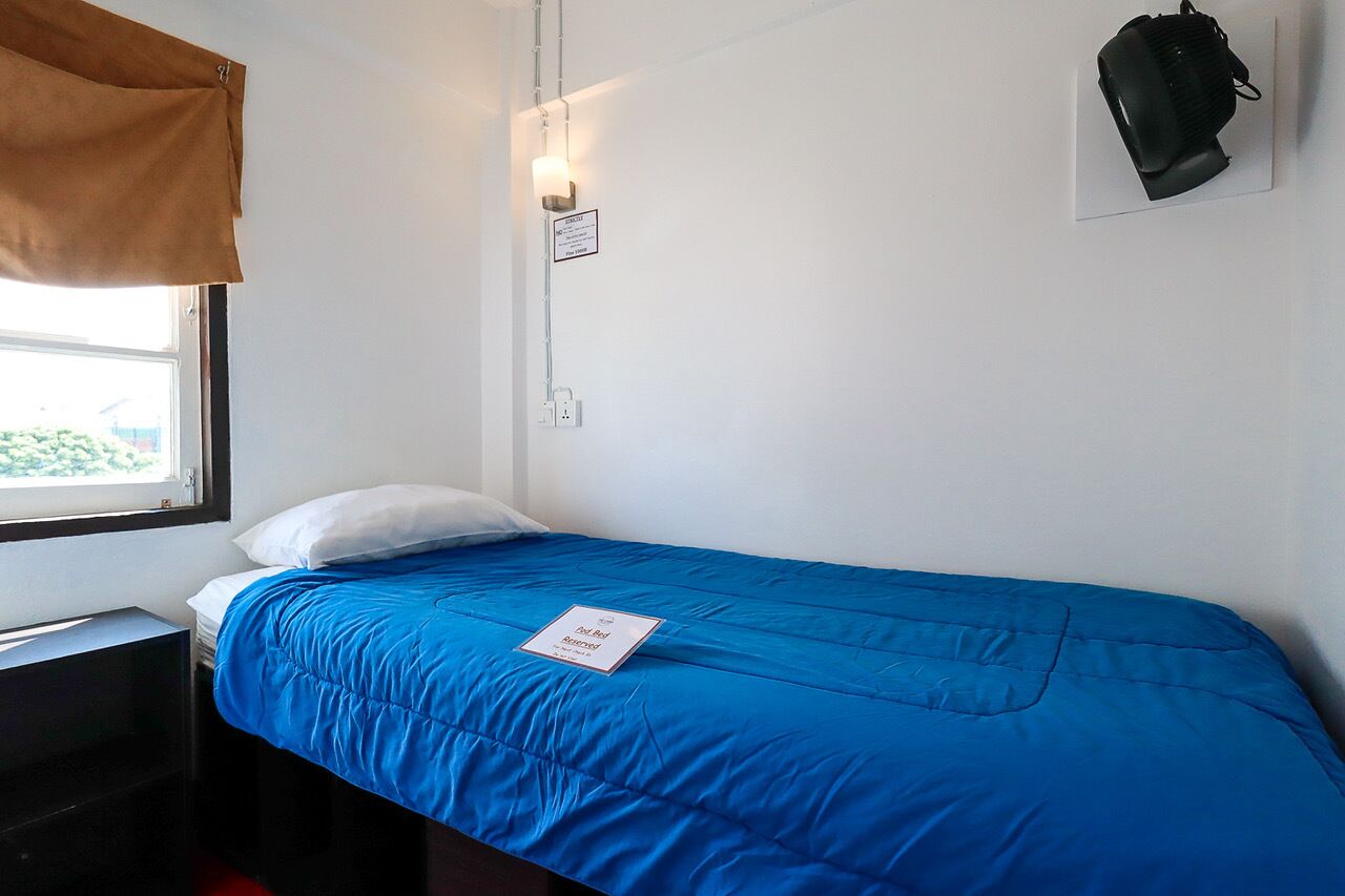 Una cama individual con colcha azul, una almohada blanca, una mochila negra en la pared y una ventana con cortina marrón.