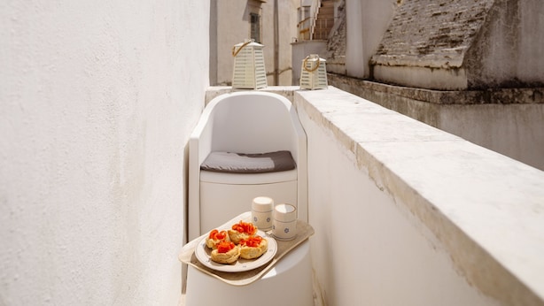 Un balcón estrecho con una silla blanca, una mesita con pasteles y una pared blanca.