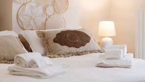 Una cama bien tendida con un cojín decorativo, toallas enrolladas y una lámpara de noche.