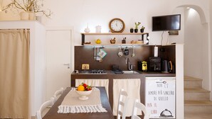 Un comedor con una mesa de madera y sillas blancas, una cocina al fondo, y un televisor montado en la pared.