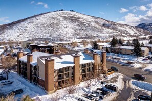 Un edificio de varios pisos con techo nevado, estacionamiento y montañas al fondo.