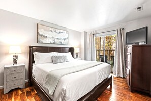 Ein Schlafzimmer mit einem großen Bett, einem Fernseher, einer Kommode und einem Fenster mit Vorhängen.