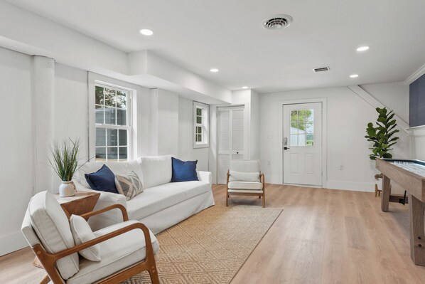 A spacious living room with a white sofa, a wooden chair, and a coffee table. There is a large window with blinds, a door, and a plant on the right side.