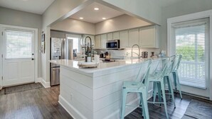 A kitchen with a large island, white cabinetry, stainless steel appliances, and a bar area with chairs. - "Bleu Fin Cottage" BHI Club, Ocean Front, Steps to Beach, Sleeps 6 (Bald Head Island)