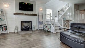 A living room with a fireplace, a staircase, a leather sofa, and a TV mounted on the wall. - "Bleu Fin Cottage" BHI Club, Ocean Front, Steps to Beach, Sleeps 6 (Bald Head Island)