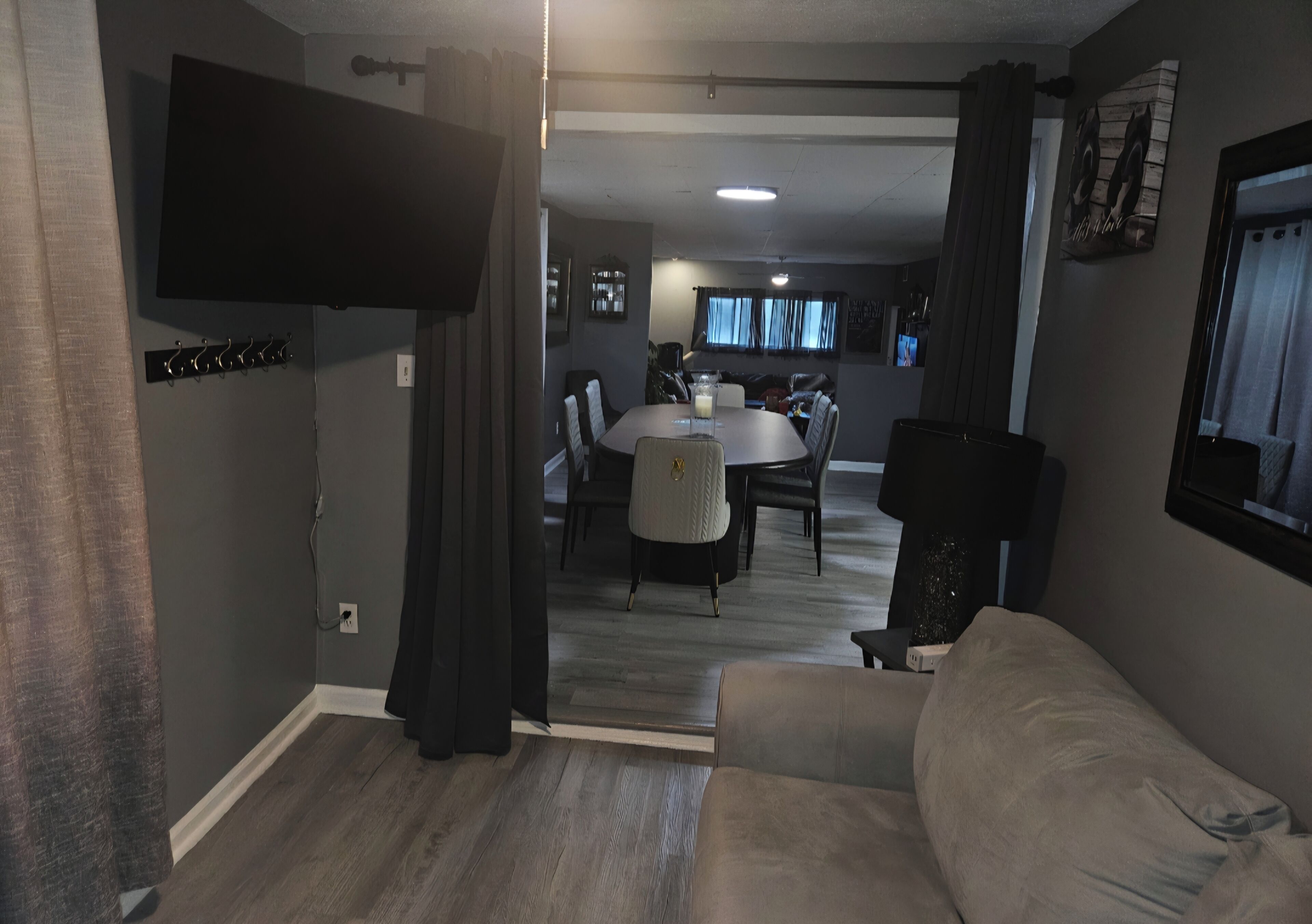 Uma sala de estar moderna com um sofá cinzento, uma área de jantar com mesa redonda e cadeiras, e uma televisão de tela plana fixada na parede.