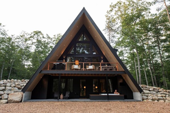 A modern, triangular-shaped cabin with a balcony, large windows, and a stone foundation.