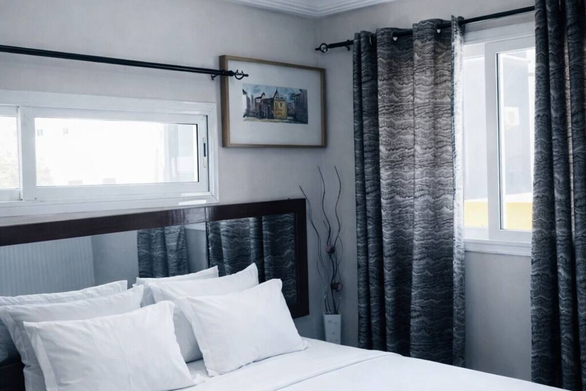 A bedroom with a bed, white bedding, a framed picture on the wall, and a window with curtains.