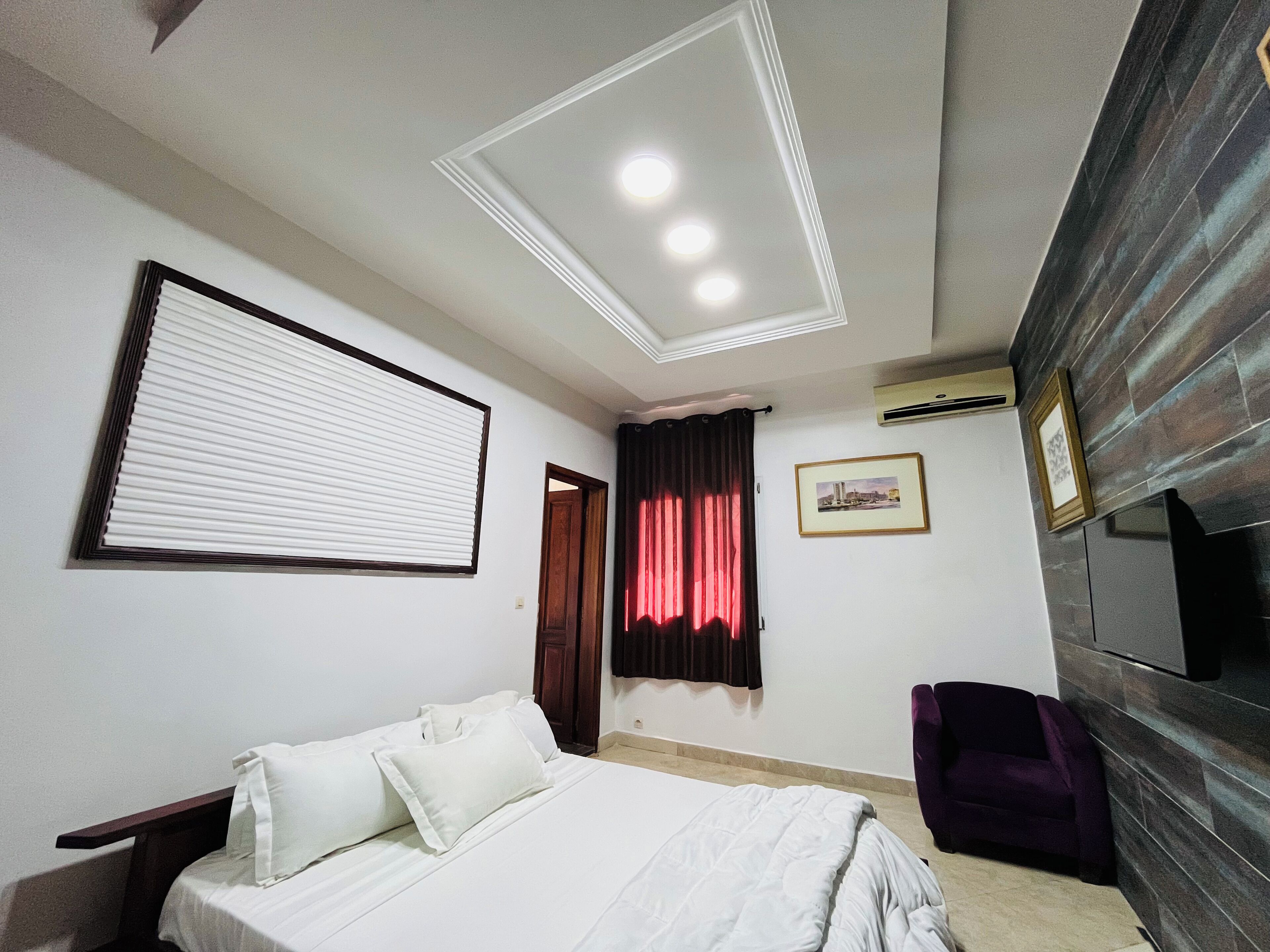 A bedroom with a bed, a window with blinds, a door, a framed picture on the wall, and a purple chair.