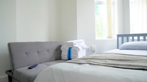 A bedroom with a grey upholstered headboard, a bed with white bedding, a remote control on a small table, and a stack of white towels with a blue ribbon tied around them.