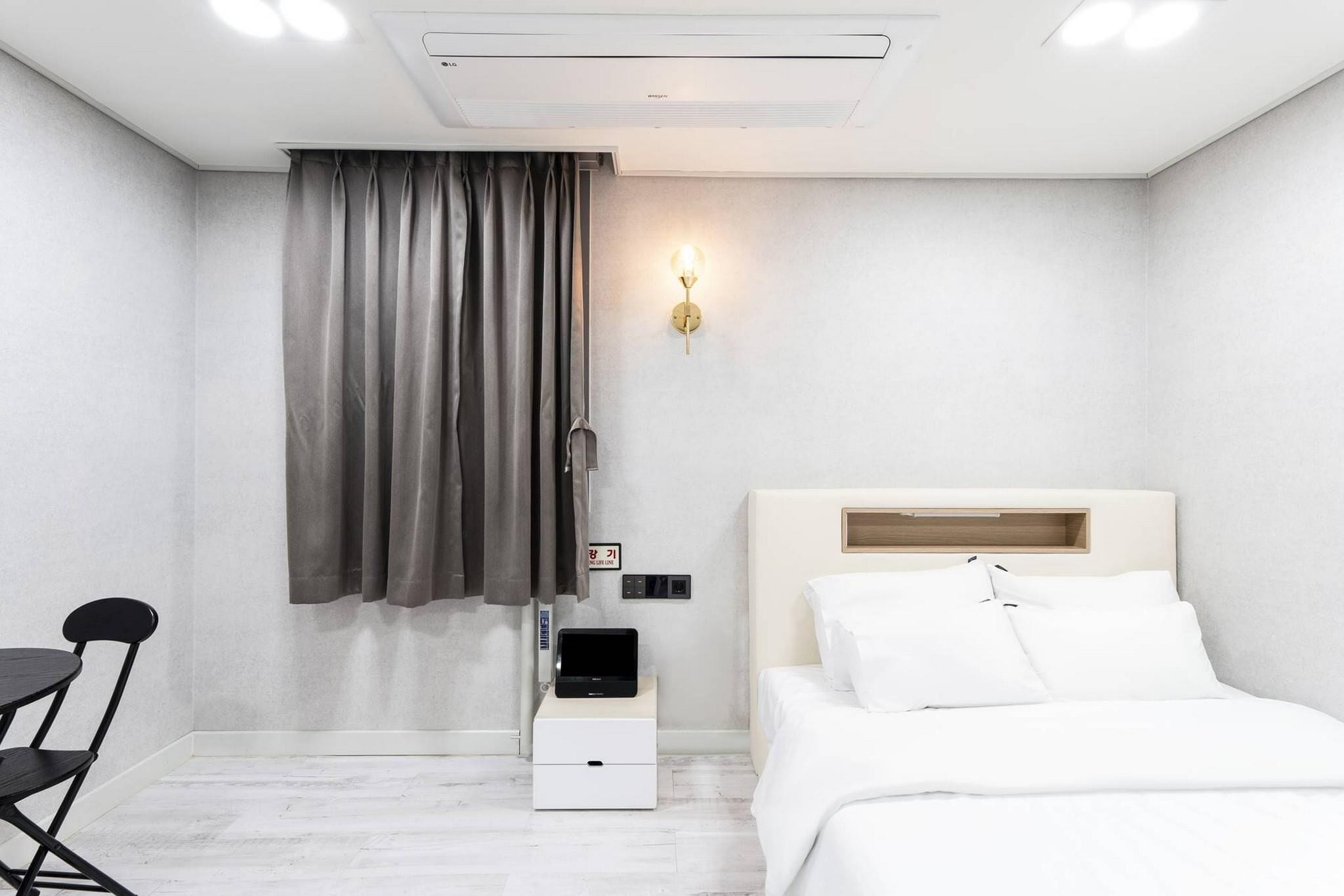 A modern hotel room with a bed, a bedside table with a lamp, a chair, and a small desk with a computer.