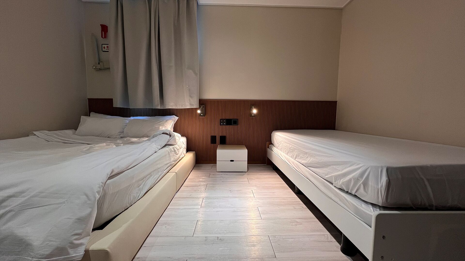 Quarto com duas camas, uma mesa de cabeceira, aquecedor fixado na parede e um armário pequeno.