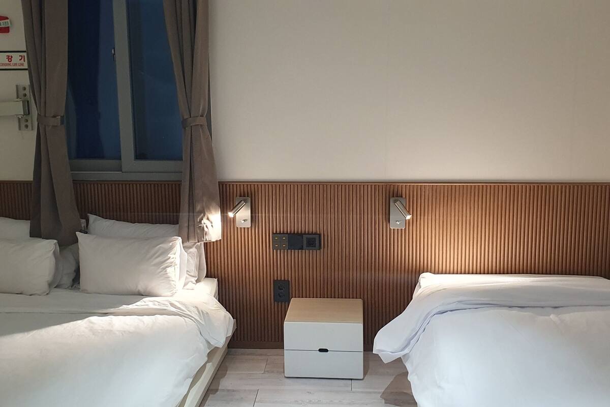 A hotel room with two beds, a wooden headboard, a window with curtains, a bedside table, and a wall-mounted emergency light.