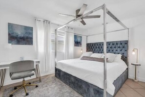 A four-poster bed with a tufted headboard, a desk with a chair, and a standing lamp.