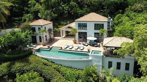 A two-story house with a swimming pool, surrounded by lush greenery.