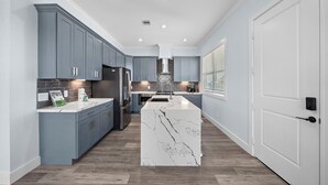 A modern kitchen with grey cabinets, a central island, and a stainless steel refrigerator. - Beachside Serenity | Modern, Near the Beach (Galveston)