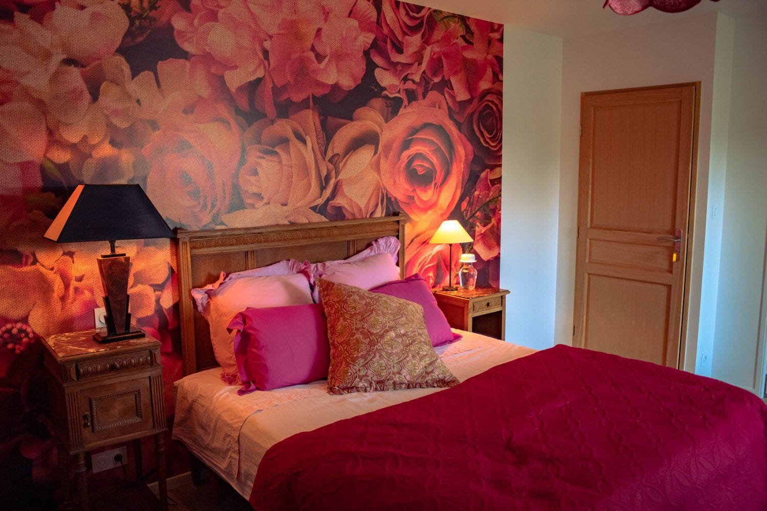 A bedroom with a large floral wallpaper, a wooden headboard, a red bedspread, and two bedside tables with lamps.
