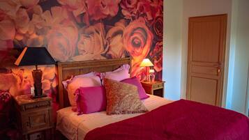 A bedroom with a large floral wallpaper, a wooden headboard, a red bedspread, and two bedside tables with lamps.