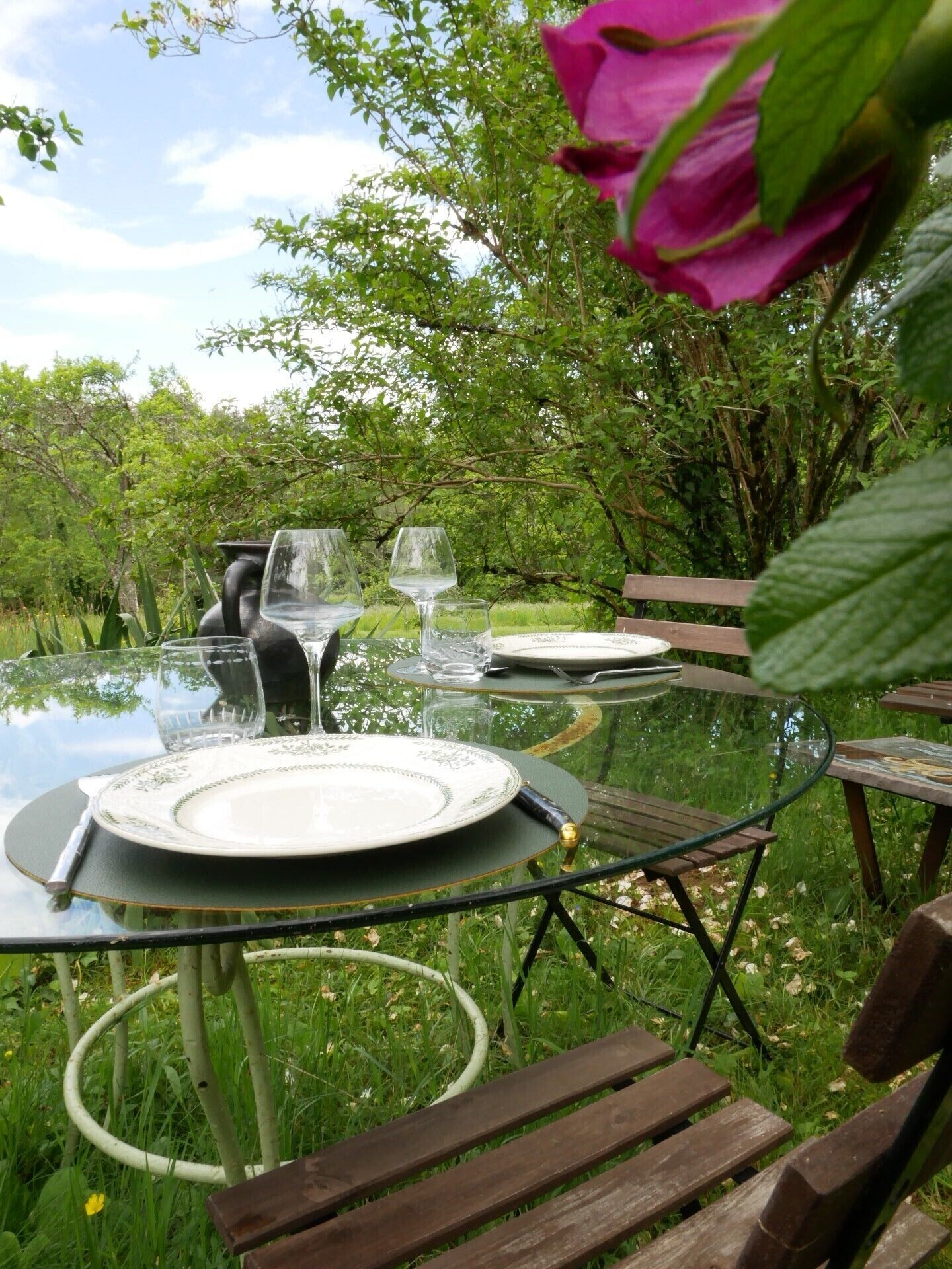 Une table à manger d’extérieur avec un plateau en verre, entourée de verdure et agrémentée d’une fleur rose.